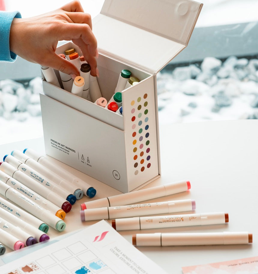 A hand taking an alcohol marker from an open box, with markers laid out on the table