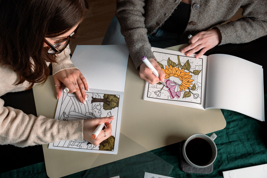 Two people coloring hand-illustrated pages with alcohol markers at a table, with coffee nearby
